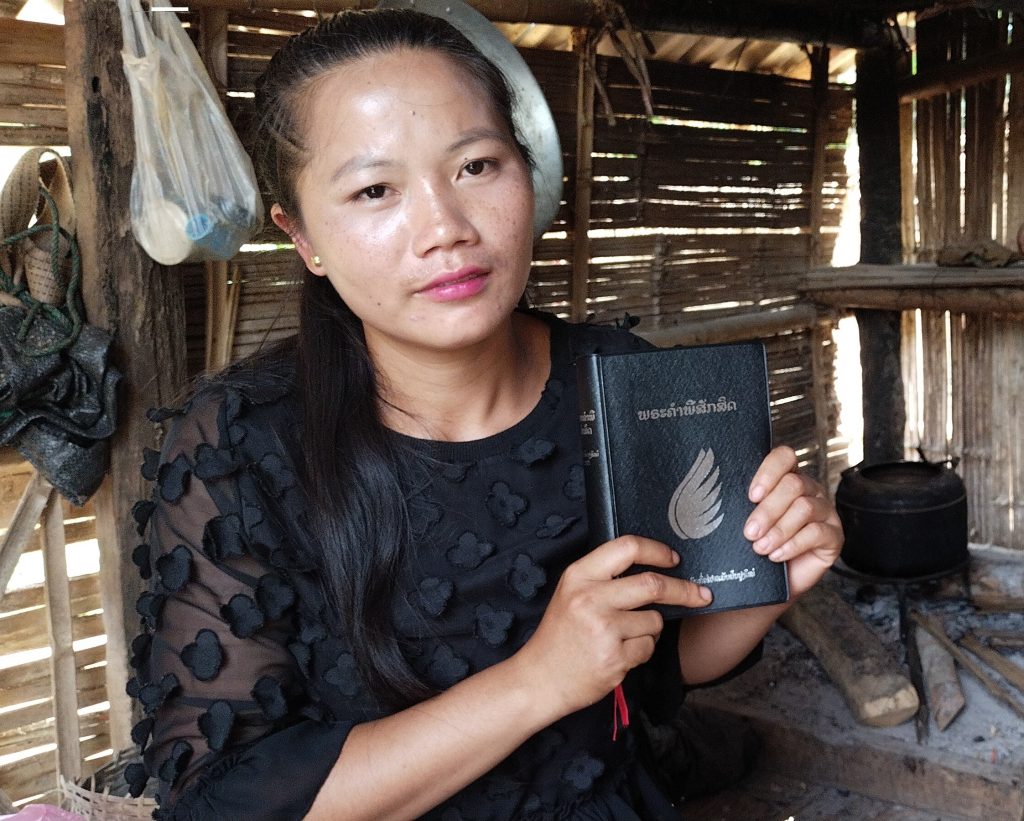 Een vrouw uit Laos houdt haar Bijbel naast zich. Op de achtergrond is haar huis zichtbaar.