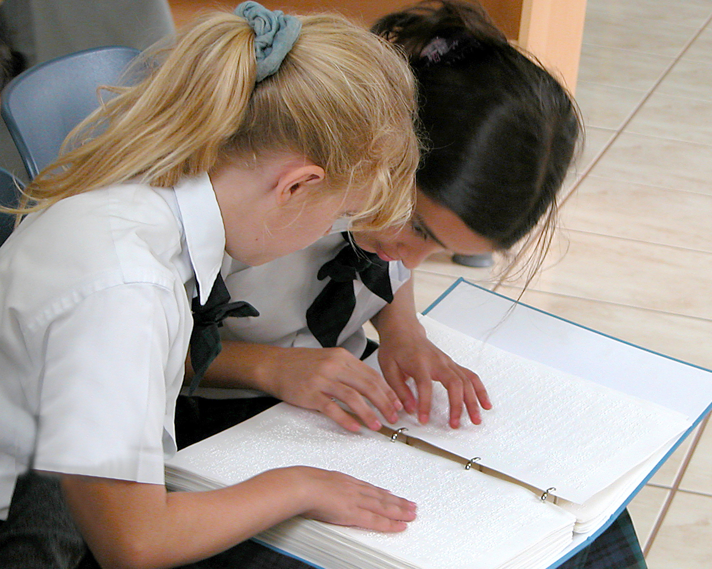 Twee meisjes zitten samen. Hun handen gaan over de bladzijden van een braillebijbel.