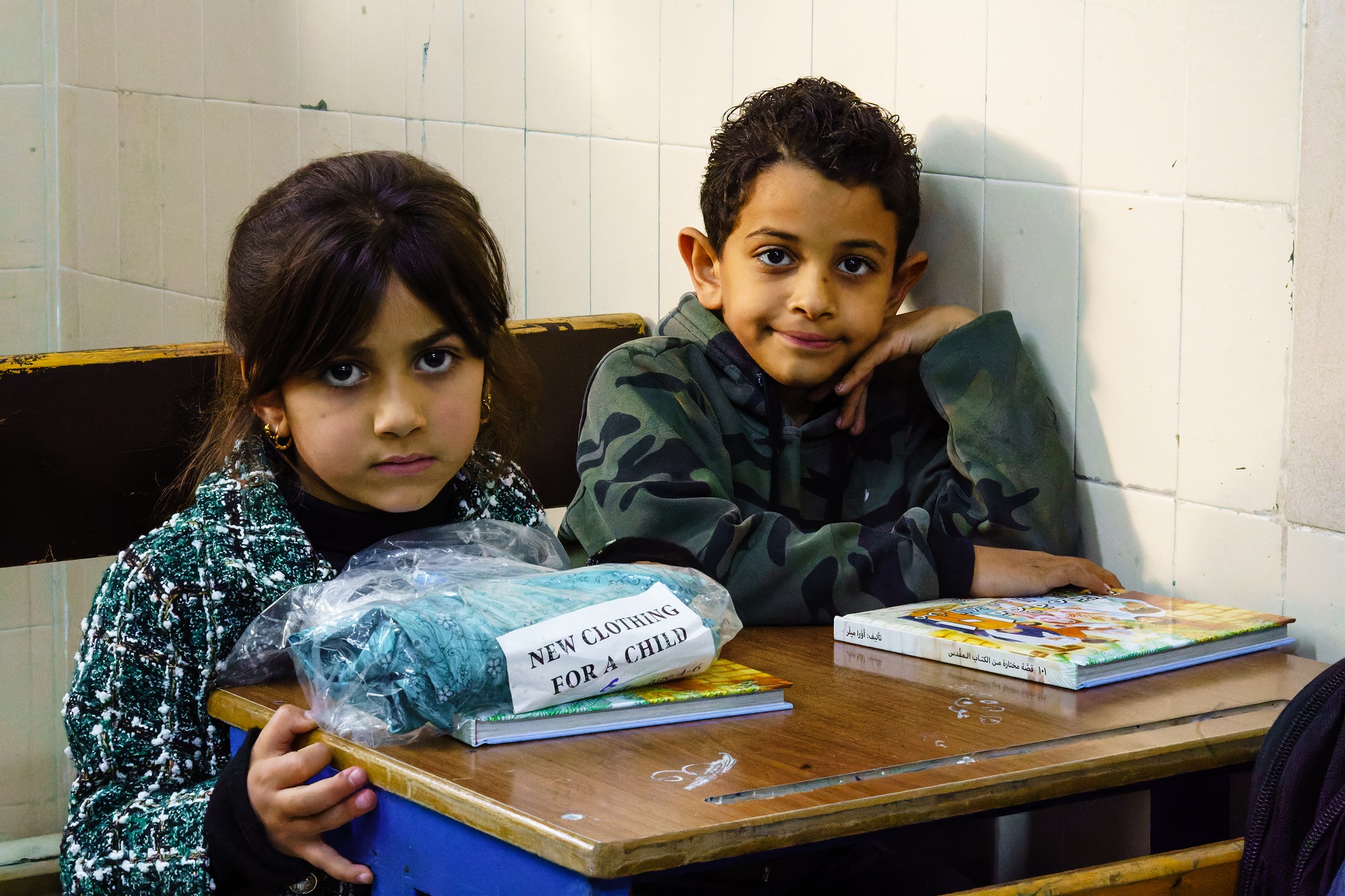 Een meisje en een jongen zitten in een schoolbankje. Voor het meisje ligt een kinderbijbel en een plastic zak met kleding. Voor de jongen ligt een kinderbijbel.